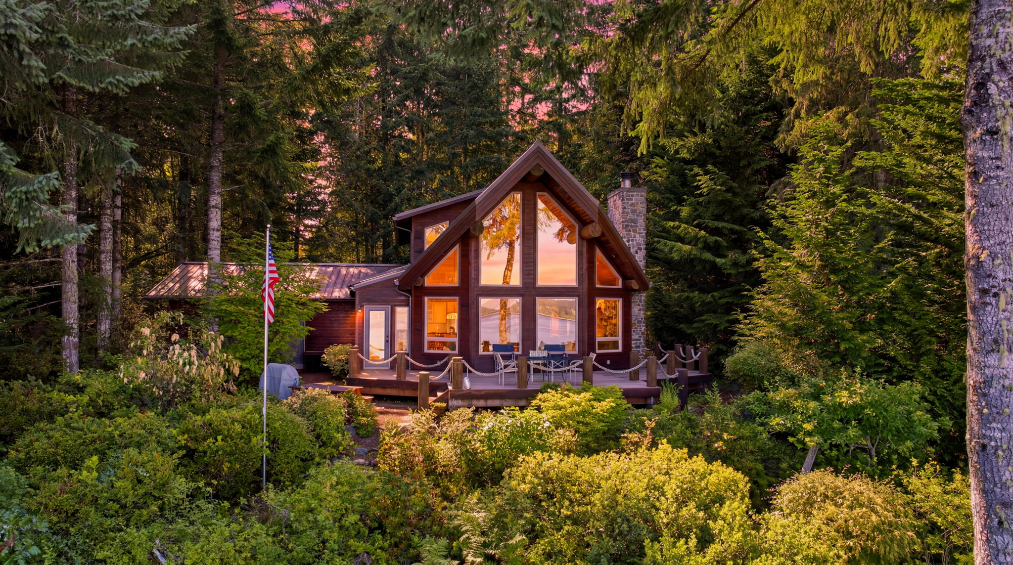A two story wood home with a steep roof and warm lights on at dusk, with a green lawn and garden in front
