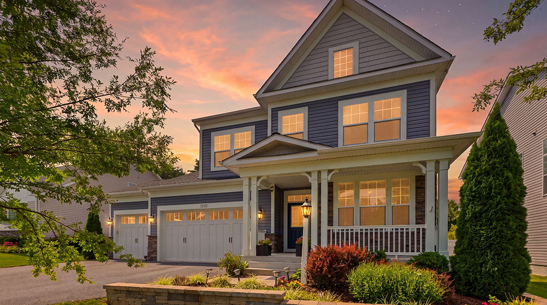 A two story wood home with a steep roof and warm lights on at dusk, with a green lawn and garden in front